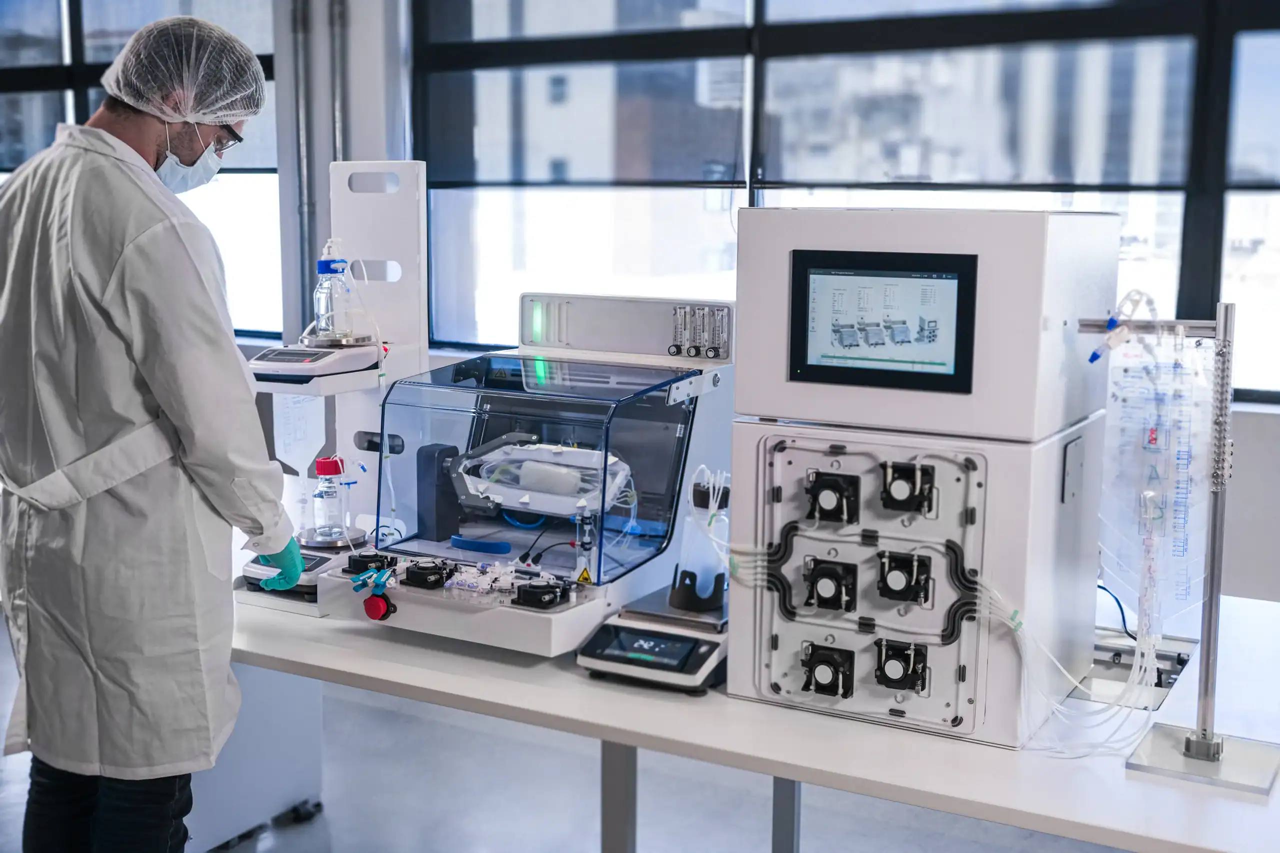 A scientist in a lab coat and hairnet operates an automated biotechnology system with a bioreactor, digital interface, and peristaltic pumps on a white laboratory bench.